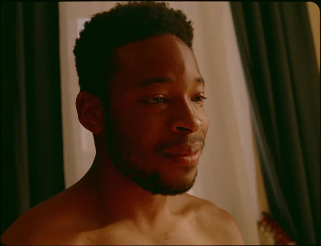 A bare chested man in his living room sits up and laughs