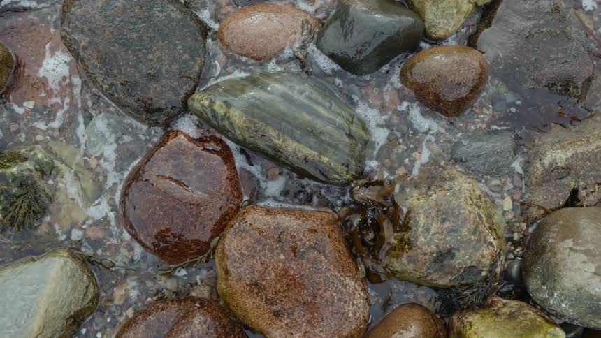 View from above of ocean waves hitting some boulders and some kelp on the beach creating seafoam