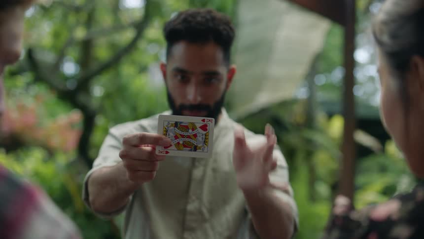 Young bearded magician entertains friends with card tricks in the garden