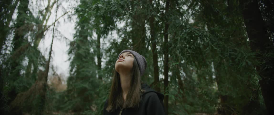 A teenage girl with long hair wearing a knit hat is standing in the woods looking up at trees
