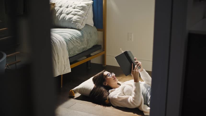 A woman is lying on her bedroom floor holding up a book as she reads