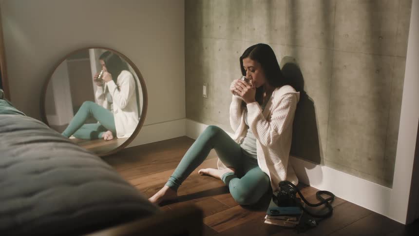 A woman is sitting on her bedroom floor leaning against the wall as she drinks from a glass and her reflection is in the mirror