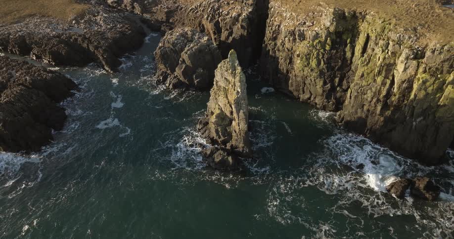 Waves splash against tall rock formations in the water near a cliff along the coast