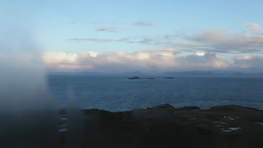 A rocky area with fog coming off of it meets the ocean with a blue sky in the background
