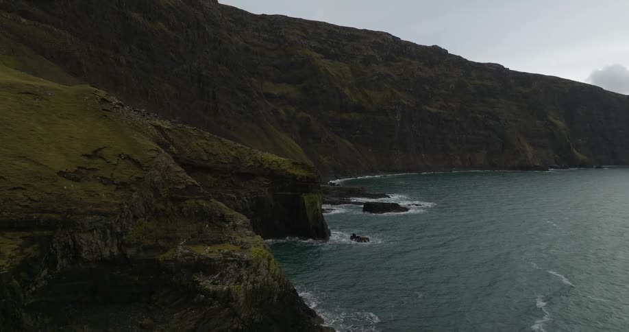 A mountain cliff covered in mossy grass towers over the rocky coastline below
