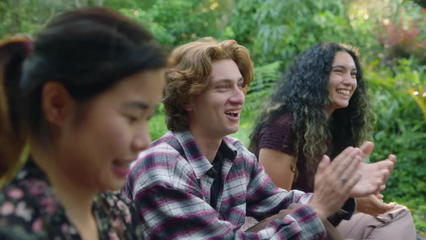 Friends sitting on ground in garden smile and clap after one of them catches playing card