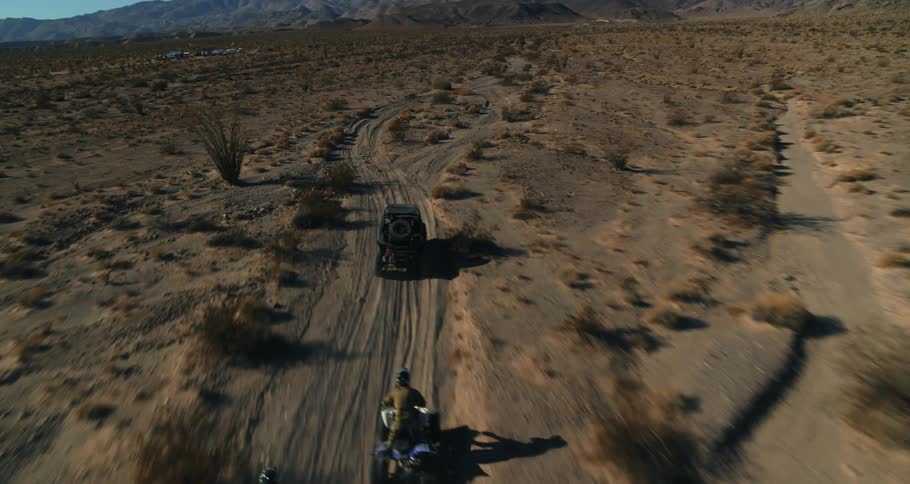 Two people riding four wheelers follow dune buggy along dirt road through arid scrub land