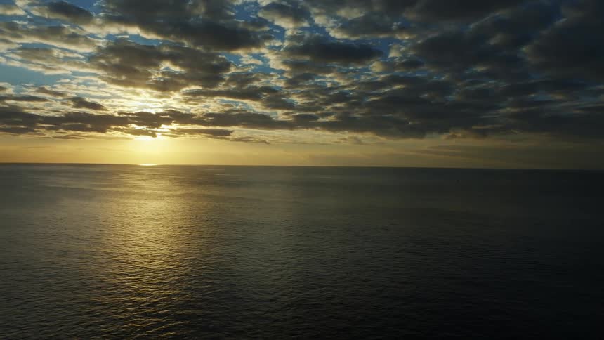 The sun shines brightly on the horizon reflecting on the rippling water under a cloudy sky