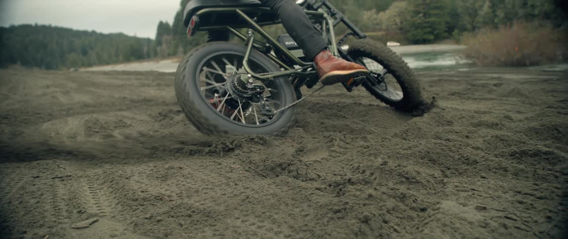 A cyclist on a bike spins the bike and kicks up dirt as they ride near a small lake