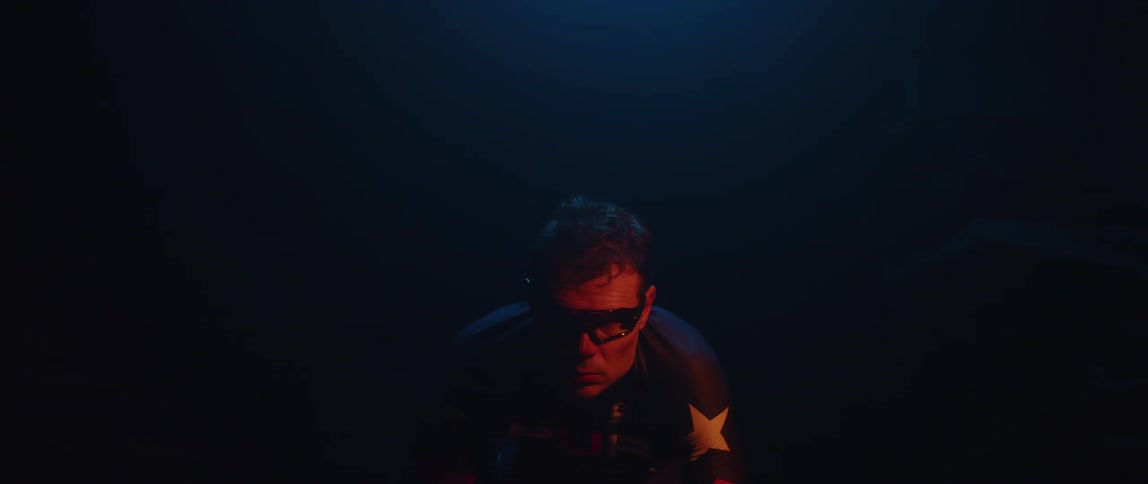 Twisting footage of a man pedaling on a static bicycle inside a dimly lit room against a dark background