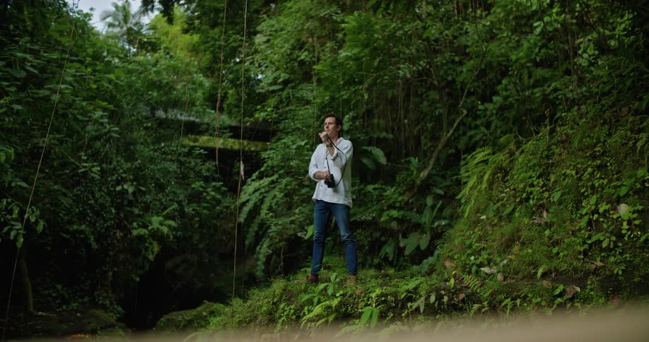 A man walks out of the rainforest and stands on a ledge as he moves the camera from his shoulder and takes a picture