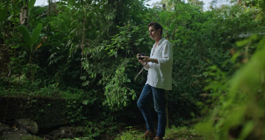A man is holding the camera strap over his shoulder as he walks through the rainforest and raises his camera to take a picture