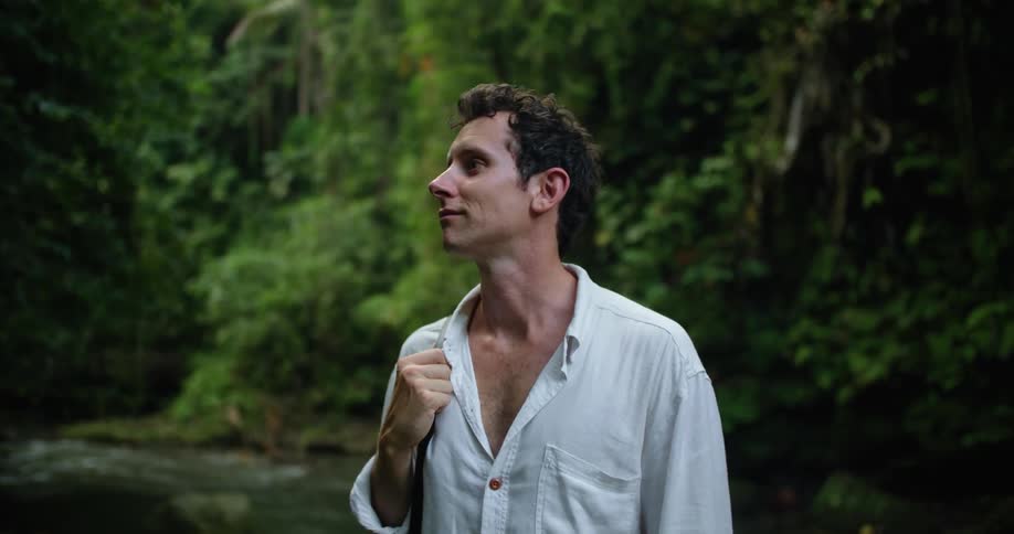 A man is standing by a stream holding his camera strap and looking up at the trees in the rainforest
