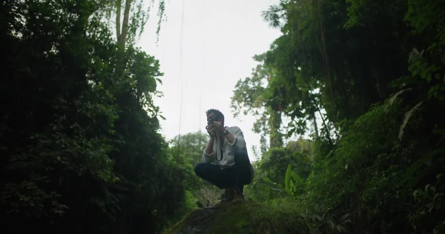 A man crouching on a boulder while photographing a jungle with a camera