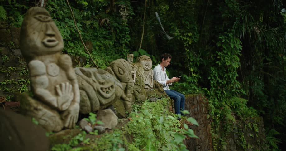 A man checks his smartphone while sitting beside a row of statues on a ledge in the wilderness during the day