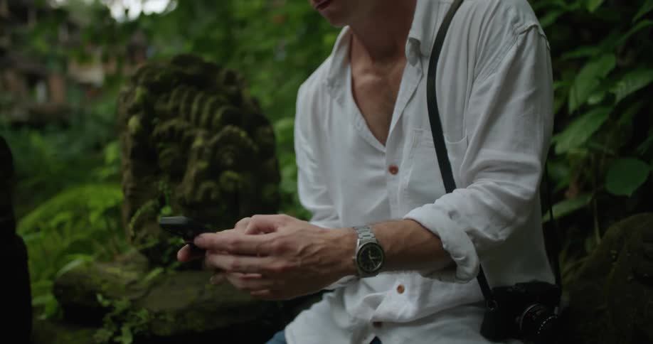 A man is sitting outside with a camera over his shoulder as he texts on his cell phone
