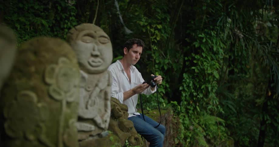 A man is sitting on a wall next to tiki statues as he lifts his camera to his face and takes a picture
