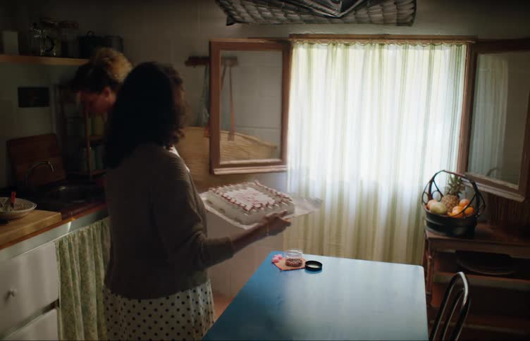 A woman at home puts a decoration on a cake while another woman walks to the sink and they both turn to leave at the same time and almost bump in to one another