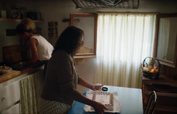 A woman decorates a birthday cake inside a house and another woman walks in and goes to the sink they both then turn around at the same time and bump in to one another