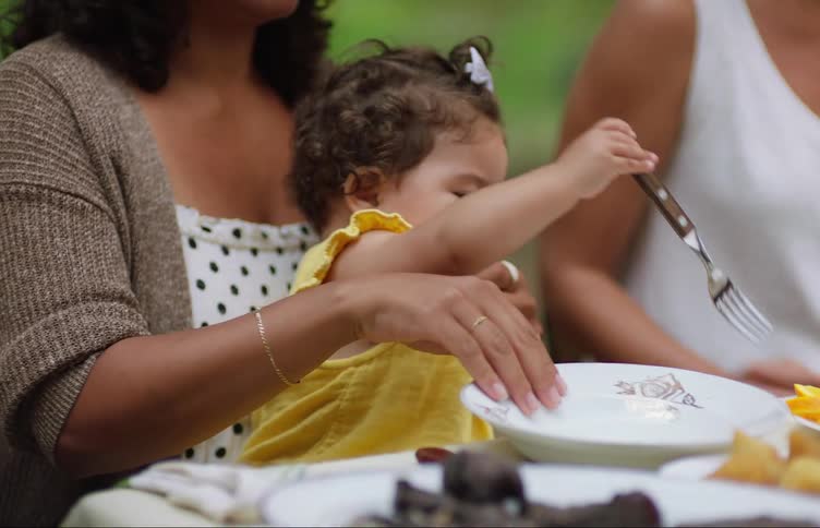 Toddler sitting on her mothers lap playing with cutlery on a table outdoors