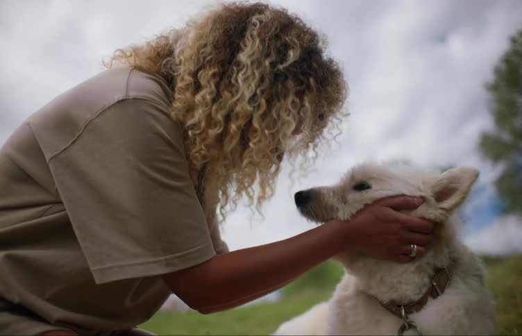 A woman with curly hair pets and scratches her white dog as they sit in a field