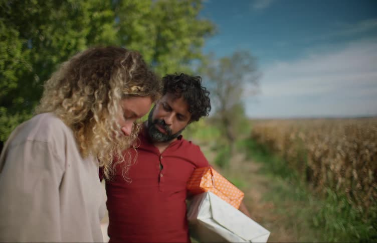 A man holding wrapped gifts stands in a field and talks and hugs a woman