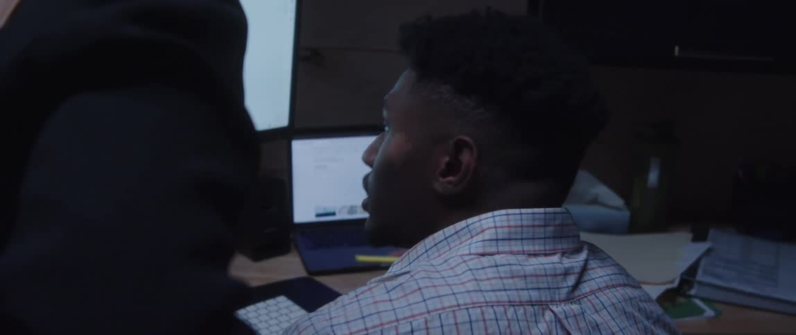 A man is sitting at a desk with a laptop computer and a monitor as he holds a pen in his hand and speaks to a woman standing next to him looking at the monitor