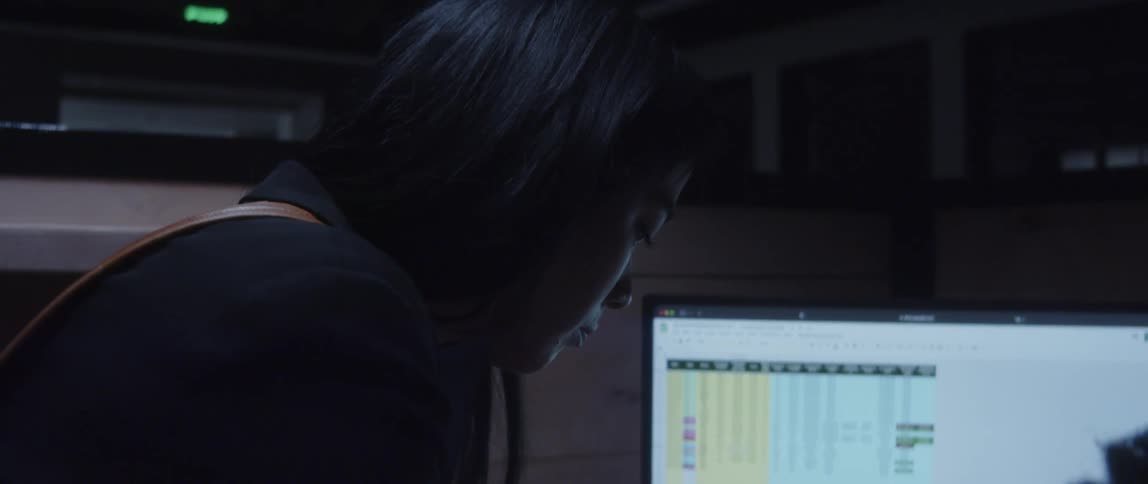 A man in an office sits in front of a computer as a woman shows him a number on a calculator