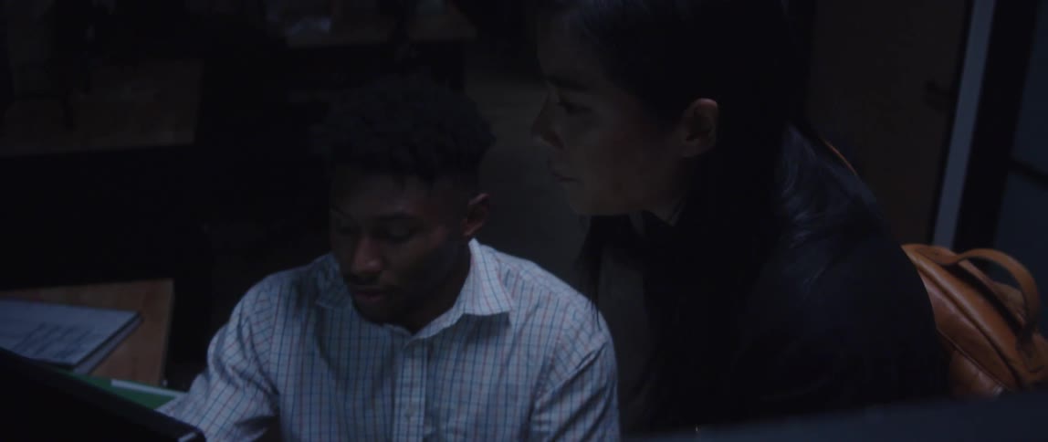 A man in an office sits at a desk and talks with a woman as they look at a computer