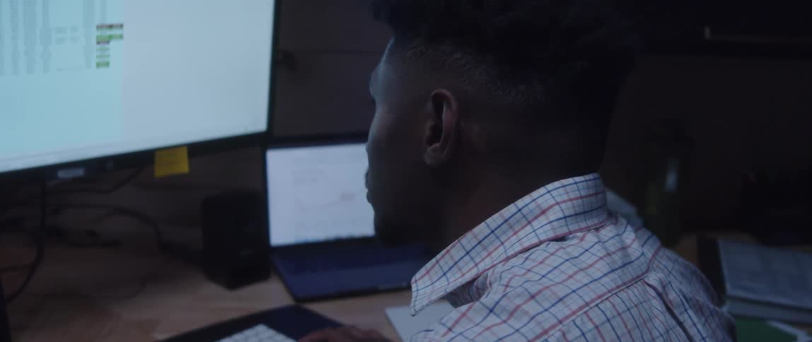 A man is sitting at a desk typing on the keyboard and looking at a large monitor with a laptop computer next to him