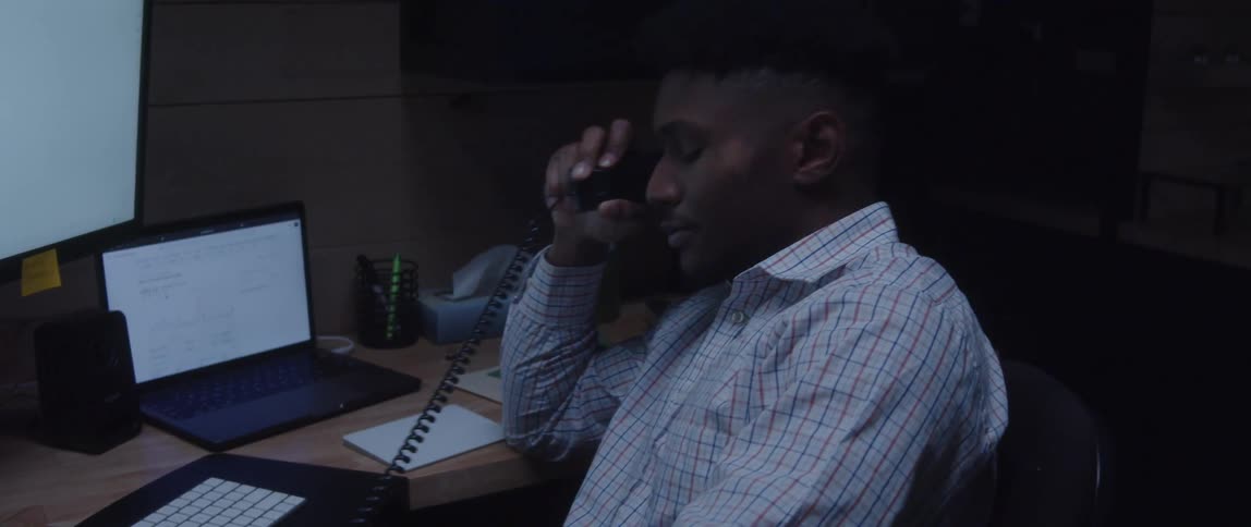 A man is sitting at a desk with computers as he talks on the telephone
