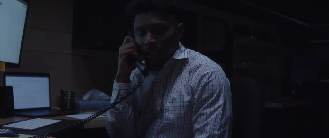 A man is sitting at a desk with computers as he talks on the telephone and leans back in his chair