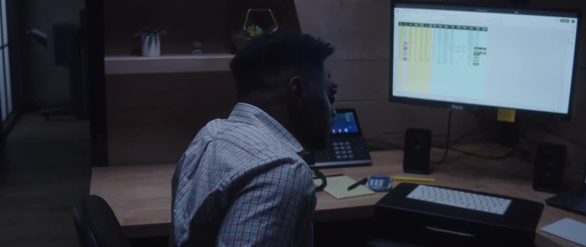 Black man sitting at desk in small office cubicle talking on landline hanging up and looking frustrated and defeated