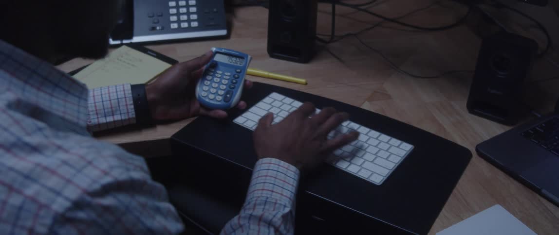 A man is sitting at a desk using a small portable calculator and looking at a chart on a computer monitor