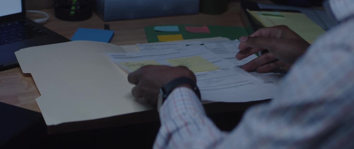 A man is sitting at a desk with computers as he looks through papers in a folder