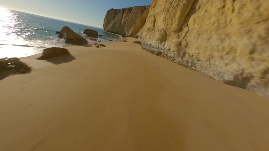 Rocks are scattered along the beach near a cliff with a cave as waves roll into shore and the reflection of the sun sparkles on the water