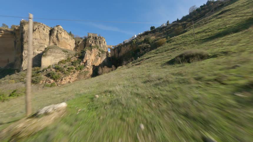 Moving across a grass covered hill with mountain cliffs ahead and buildings overlooking the valley and arches made of stone