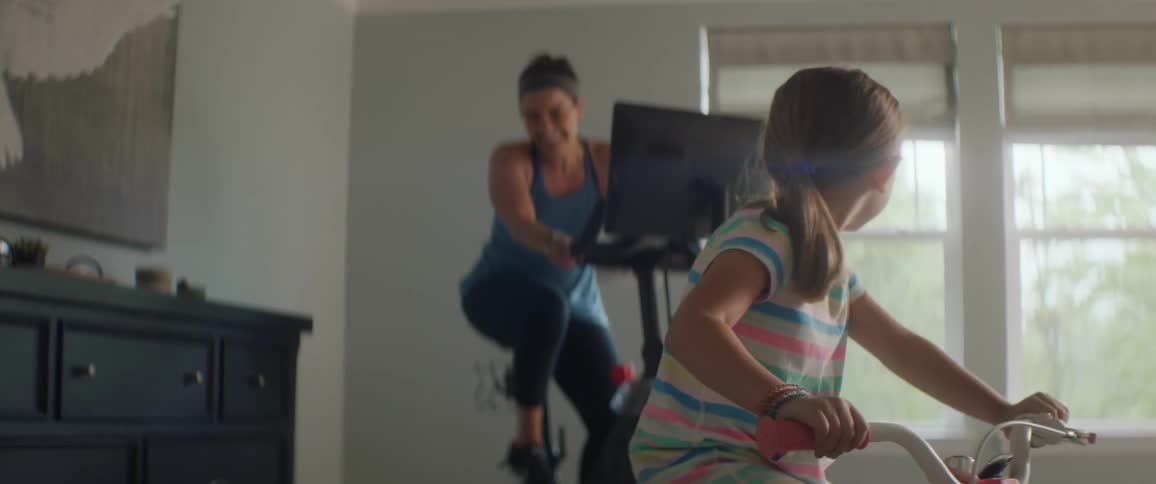 A young girl rides a stationary bike indoors and her mother rides an exercise bike behind her