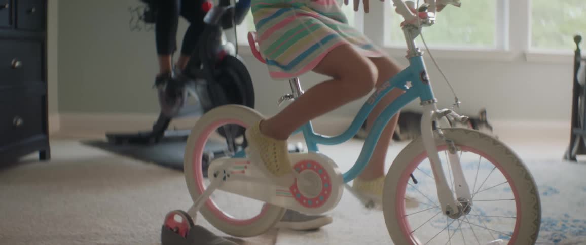 A woman users her exercise bike and a girl uses her bicycle in a bedroom