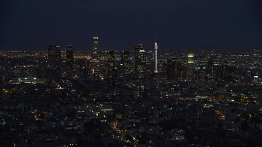 Slowly approach the towering skyscrapers of Downtown Los Angeles in a nighttime aerial clip during a partly cloudy evening