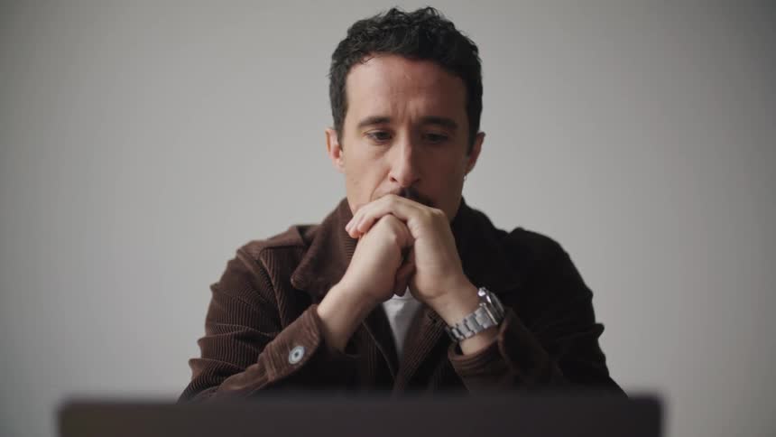 A man sits in front of a plain white wall typing on the computer in front of him thinking and looking away while placing his hand together under his chin dismayed