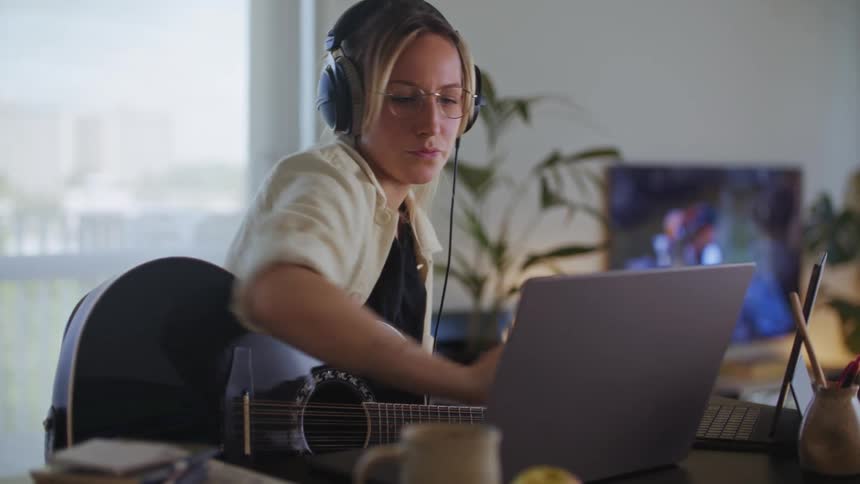 A woman sitting at a desk wearing headphones and playing an acoustic guitar is working with two laptop computers