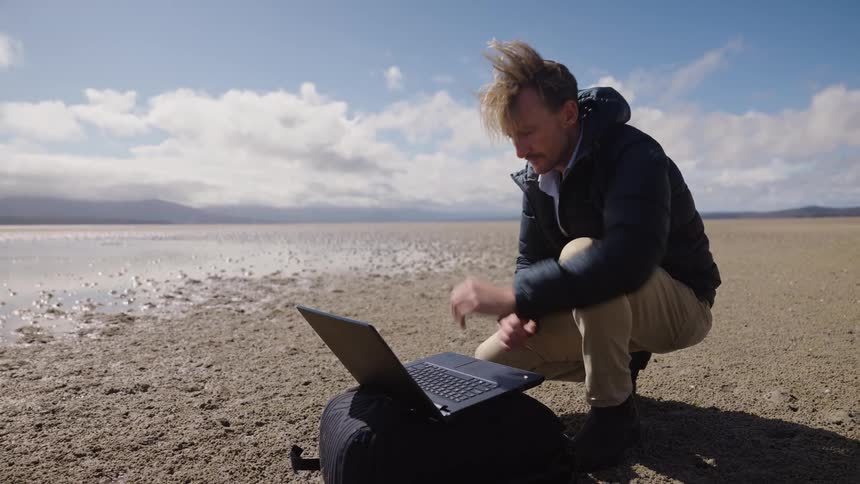 A man kneeling in front of his laptop sitting on a bag on the ground as he looks around the desert he is in and types on his laptop