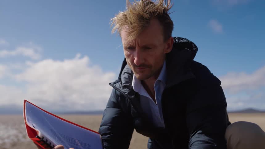 A man in a warm coat is bent down on one knee typing with one hand on a laptop computer that is sitting on top of a backpack with a notebook in the other hand alone on a sandy beach