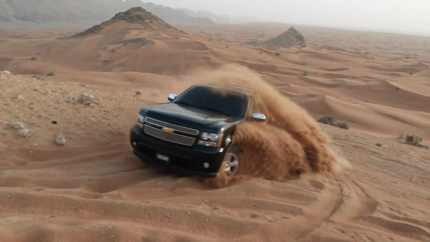 A black truck speeds around a curve as it travels up a desert hill and sand flies as it moves