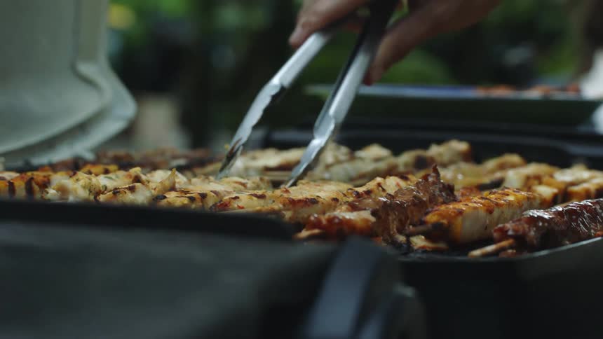 A hand is holding a pair of tongs flipping meat on sticks over on an outdoor grill beside a plate sitting on top of the grill side table and beyond that a person is sitting down in front of green trees or shurbs