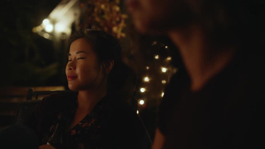 Two people are sitting down outside at night while one has her eyes closed and is slowly moving her head back and forth