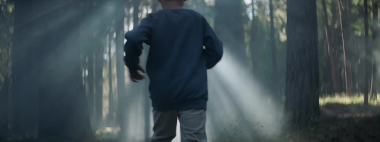 A little boy runs through the forest as rays of sunlight shine through the trees