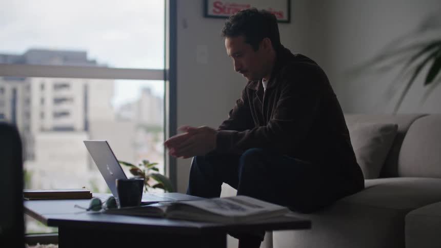 A man is sitting in front of a desk as he opens his laptop computer and types