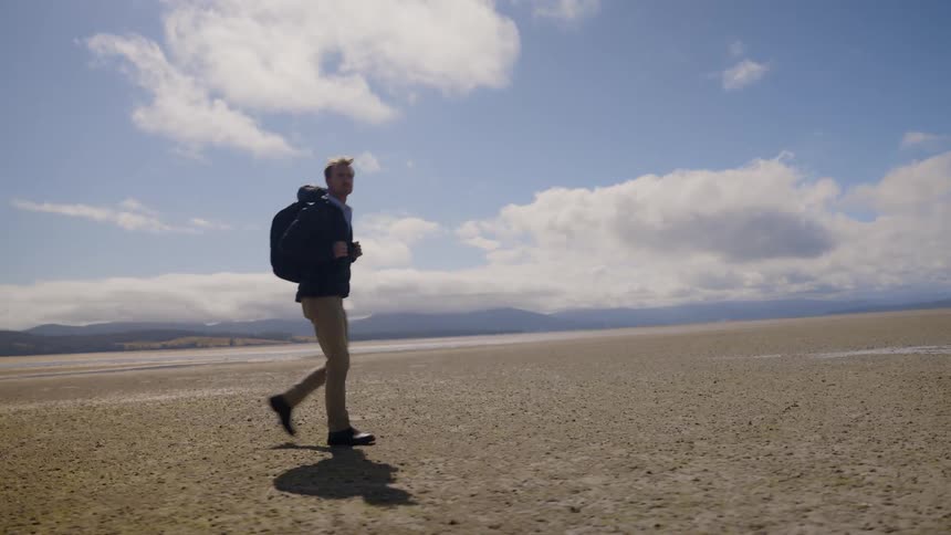 A man wearing a jacket and a backpack is walking across the desert and there are mountains in the distance under a cloudy sky